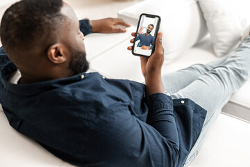 Two diverse male friends or colleagues talking on the distance. African-American man lying on white sofa in modern apartment, using smartphone for video call, talking with indian guy