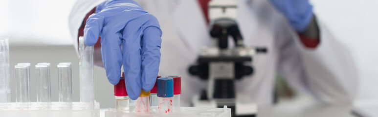 cropped view of scientist in latex gloves reaching test tubes near blurred microscope, banner.