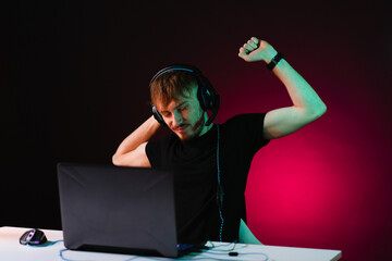 Portrait of Software Developer having a break. Hacker. Gamer Wearing Headset Sitting at His Desk and Working. Playing on Laptop. In the Background Dark Environment with red neon lights.