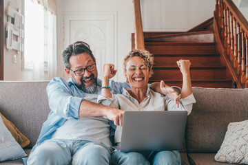 Astonished caucasian couple celebrating online success using laptop sitting on sofa at home. Joyful...