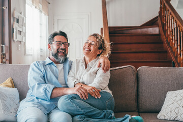Happy caucasian couple laughing while sitting on sofa in the living room. Husband and wife laughing out loud sitting on couch at home. Man and woman spending leisure time at modern apartment - obrazy, fototapety, plakaty