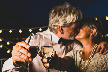 Old caucasian couple kissing each other while toasting champagne glasses celebrating new year party. Loving husband and wife kissing each other while partying with white wine glasses in hands - Powered by Adobe