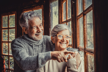 Old caucasian couple spending leisure time looking out through window at home. Loving husband...