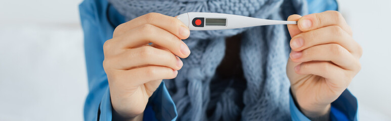 Fototapeta premium cropped view of sick woman in knitted scarf holding electronic thermometer, banner.