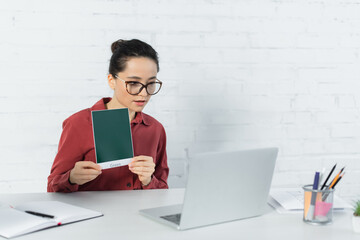 Obraz premium young teacher in eyeglasses holding card with green lettering and looking at laptop on desk.