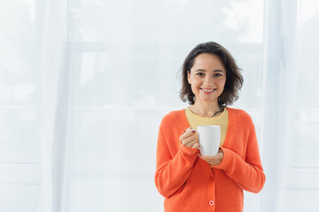 positive young woman smiling and holding cup near white curtain.