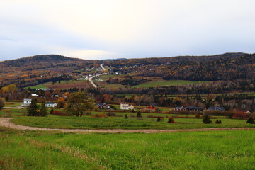 Fototapeta na wymiar View of a little village in Charlevoix. Autumn landscape in october. Autumnal. Hill and trees in auutmn. Grass and wild fleld in autumn.