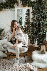 Overjoyed young happy european woman in warm knitted sweater sitting under decorated Christmas tree, holding mobile call conversation with relatives or friends, congratulating telling best wishes.