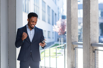 Fototapeta premium Exited African American Businessman Celebrating Achievement looking at Smartphone. Successful business man victory win with mobile phone outside. Happy Good News Success Deal, Startup Bet winning rate