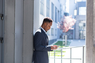 Fototapeta premium Successful and happy african american businessman near office holding mobile phone smiles and rejoices reads news and writes