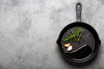 Black cast iron frying pan with ingredients for cooking: oil, fresh green rosemary, raw garlic cloves and colorful peppercorns on gray stone background flat lay top view with space for text 