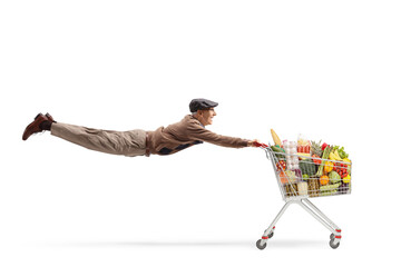 Full length shot of an elderly man flying and holding a shopping cart with food