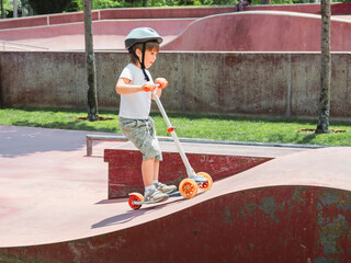 Little boy rides kick scooter in skate park. Special concrete bowl structures in urban park. Training to skate at summer.