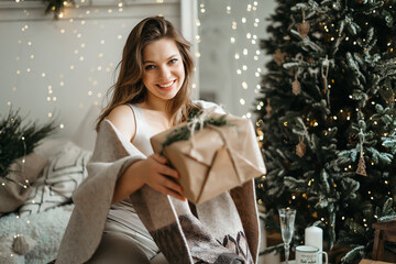 Smiling  beautiful young woman in comfortable light winter clothes holds out a gift box , prepares a Christmas present, celebrates the winter holidays