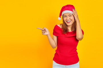 Fototapeta na wymiar Smiling young woman wearing christmas hat with open mouth holding head in hands and pointing aside