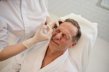 Obraz na płótnie Canvas High angle view of a senior European man receiving beauty injection on his face in wellness spa. Mesotherapy, male anti-aging procedure, rejuvenating treatment, professional skin and body care concept