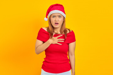 Shocked young Asian woman wearing Christmas hat and put hands chest on yellow background