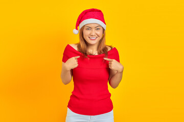 Cheerful young woman wearing Christmas hat pointing to herself on yellow background