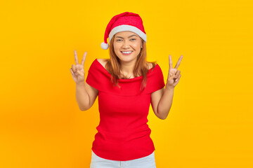 Fototapeta na wymiar Cheerful young Asian woman wearing Christmas hat showing peace sign on yellow background
