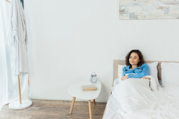 pleased woman lying in bed near bedside table with books and alarm clock.