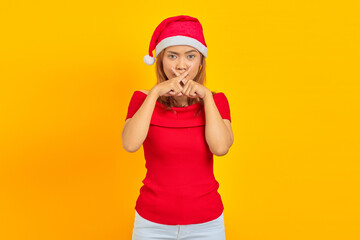 Young Asian woman wearing Christmas hat showing a sign of silent gesture on yellow background