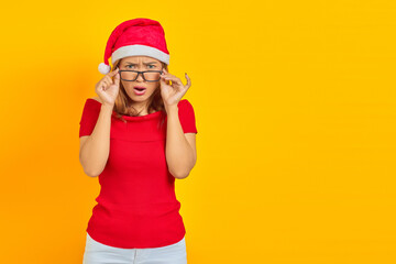 Surprised young Asian woman wearing Christmas hat with glasses while open mouth on yellow background