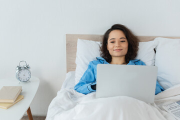 positive young freelancer in blue pajamas using laptop in bed.