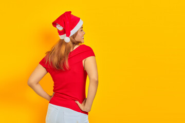 Young woman wearing Christmas hat in back position holding hands on waist confidently on yellow background