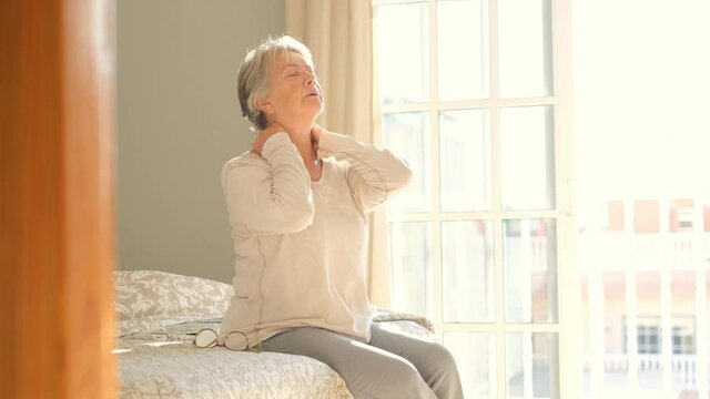 Senior woman suffering from neckache after sleep, rubbing stiff muscles, Old female sitting on bed touching neck feeling discomfort because of uncomfortable bed at home
