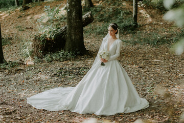 The bride in a wedding dress holds a bouquet in her hands and stands in a green forest