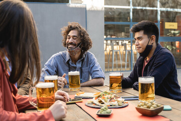 Multiracial people having fun drinking beer at brewery bar outdoor while wearing safety masks -...