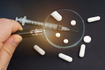 A syringe and pills lie on a black background. A person examines them through a magnifying glass.