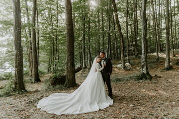 The groom in a black suit and the bride in a white dress stand and hug each other in the green forest