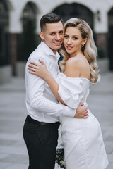 Naklejka premium A stylish, smiling groom and a beautiful, young, curly blonde bride in a white dress are embracing against the backdrop of the city and buildings. Close-up wedding photo of the newlyweds.