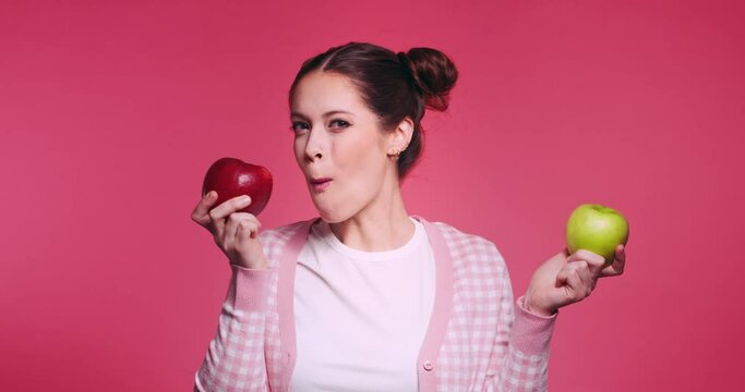 Beautiful young caucasian girl holds two apples, red and green in her hands and funny bites quickly each one in turn isolated on pibk background. Vegetarian healthy fruits. Problem of choice concept