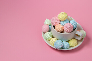 Multicolored meringues on a pink background. A teacup overflowing with colored meringues.