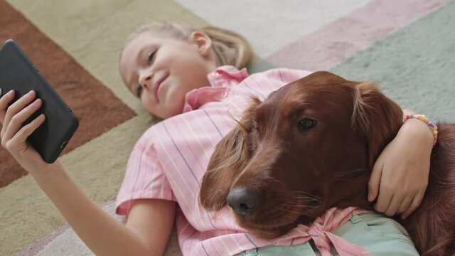 From-above medium close-up of smiling 8-year-old girl scrolling on smartphone while lying on carpet in bedroom together with her cute Irish setter dog