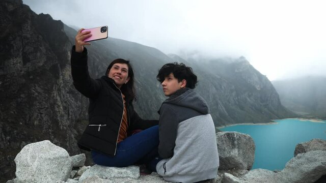 a mother and her teenage son sitting taking a selfie in an incredible background of a turquoise lagoon called Paron in Huaras with mist on the hills
