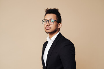 Portrait of young Afro businessman wearing shirt, black suit, glasses and looking at camera while standing at beige background.