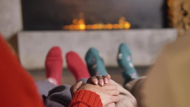 Cozy christmas atmosphere, multicultural couple are holding hands while sitting on the bed against the background of the electronic fireplace, the family shakes their legs in green and red socks from