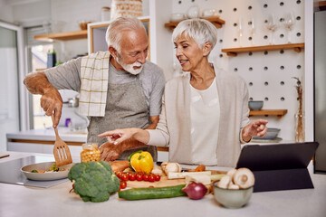 Smiling elderly vegan couple preparing a fresh vegetable salad in the kitchen, using a tablet