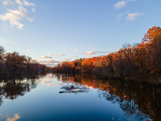 A calm lake reflects the changing leaves on the trees lining the shore, the sky touched by fluffy clouds
