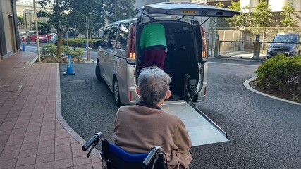 車椅子に乗った老人男性を介助しながら福祉車両に乗せる介護施設職員