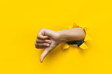 Hand showing a thumb down through ripped hole in bright yellow paper background. Concept of dislike...