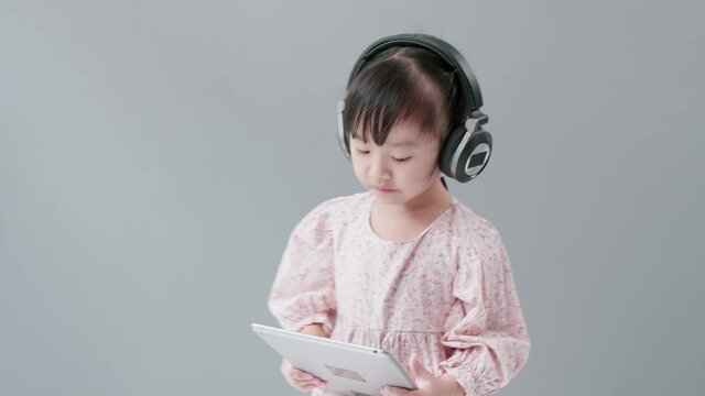 Little Asian girl wearing headphones holding tablet, listening to music and dancing happily.