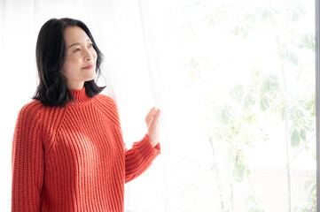 Beautiful Japanese (Asian) woman smiling as she opens the curtains and watches the sunrise in a brightly backlit room.	
