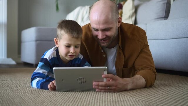 Father and son study together. Adorable little boy does interesting tasks on tablet computer spbd while bald parent helps lying on floor in living room
