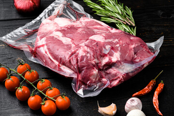 Fresh, raw, vacuum packed cut lamb meat, with ingredients and herbs, on black wooden table...