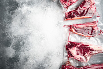 Fresh tasty raw beef steaks with bone wrapped in vacuum plastic packing at market, tomahawk, t bone, club steak, rib eye and tenderloin cuts, on gray stone background, top view flat lay
