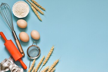 Baking cooking ingredients. Bowl with flour, eggs, eggbeater, ears of wheat, rolling pin and kitchen textile on blue background. Top view with copy space. Flat lay